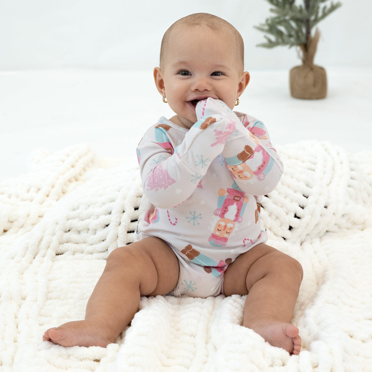 A baby sitting on a chunky white knit blanket, wearing a long-sleeve bodysuit with a colorful winter-themed print featuring snowflakes, candy canes, and nutcracker motifs.