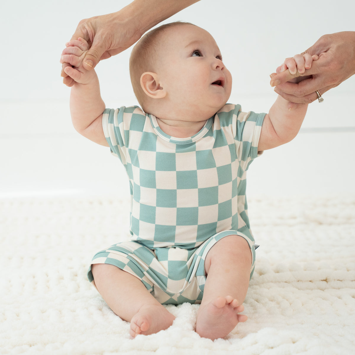 A baby wearing a mint green and cream checkered short-sleeve romper sits on a white textured blanket while holding hands with adult hands.