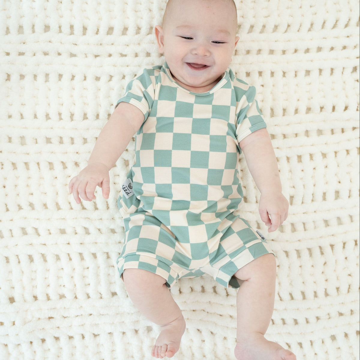 A baby lying on a white textured blanket, wearing a mint green and cream checkered short-sleeve romper.