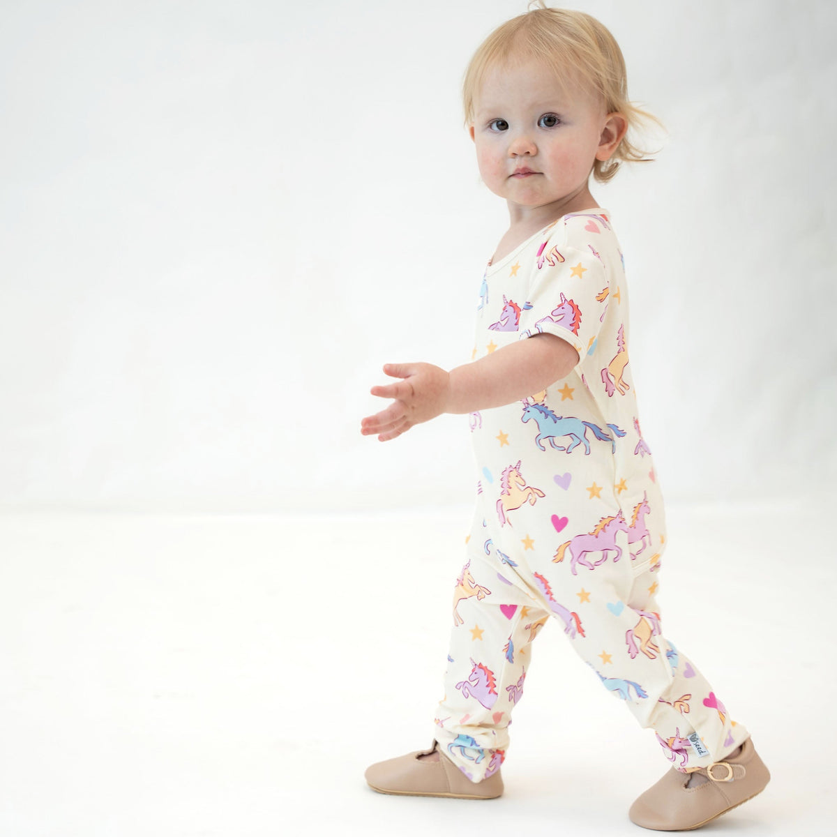 A toddler girl walking while wearing a cream-colored short-sleeve one-piece jumpsuit with a colorful unicorn print, paired with tan booties, against a white studio background.