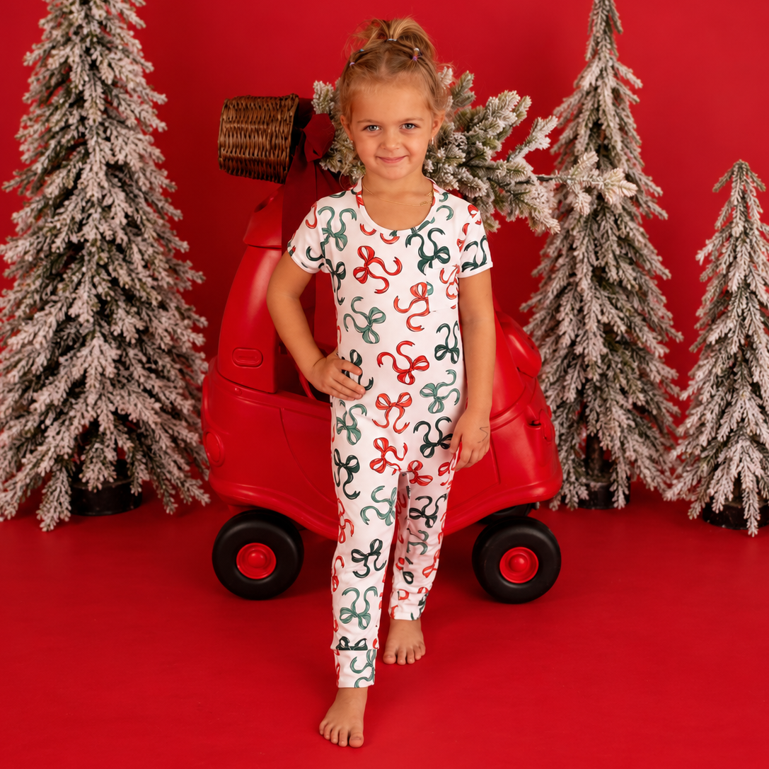 A young girl stands barefoot in front of a red backdrop, posing in a white short-sleeve pajama set printed with red, green, and black Christmas ornaments. She is posed in front of a red toy car and three flocked Christmas trees.