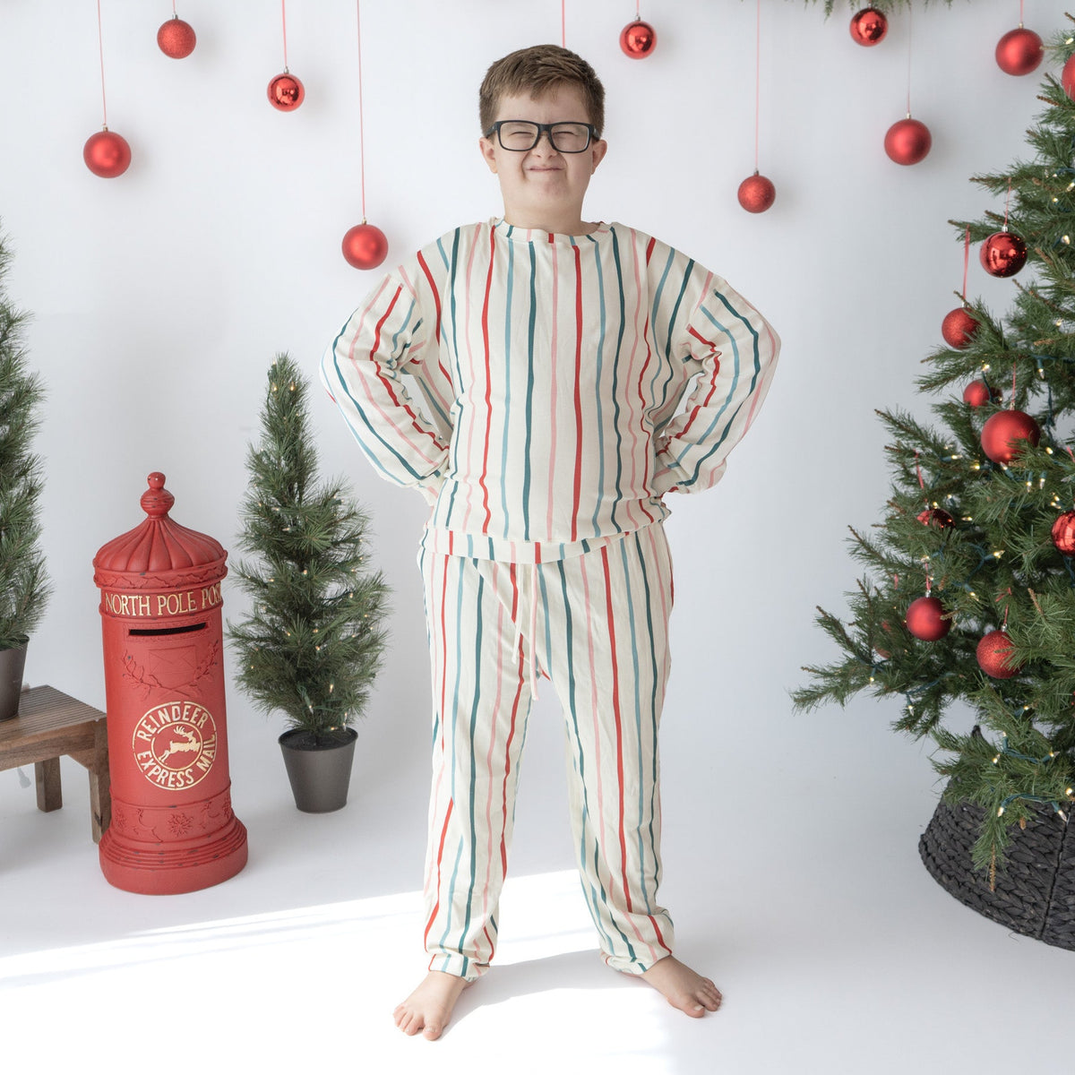 A person wearing a matching two-piece pajama set with vertical red, green, and blue stripes on an off-white background, standing in a decorated Christmas room with a tree, garland, and red ornaments.
