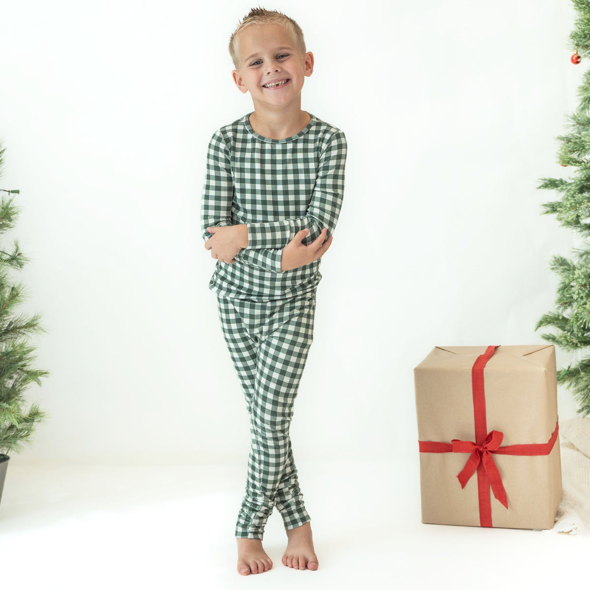 A young boy stands barefoot in a white studio setting wearing a green and white gingham long-sleeve pajama set. A wrapped gift box with a red ribbon sits to his right, and parts of a Christmas tree are visible at the edges.