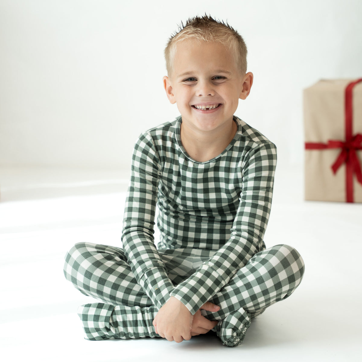 A smiling young boy sits cross-legged on a white floor wearing a green and white gingham long-sleeve pajama set, with a wrapped gift box blurred in the background.