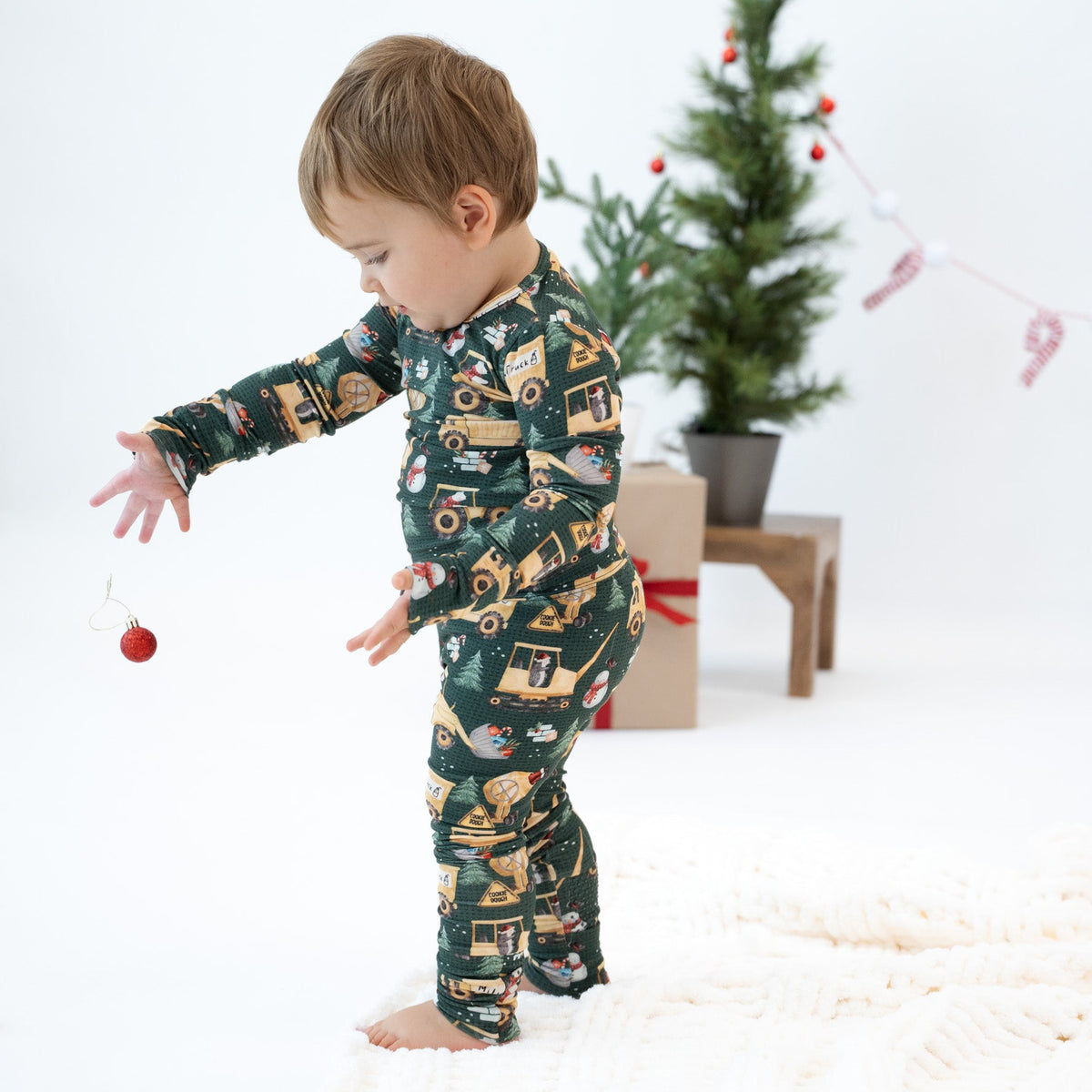 A toddler stands barefoot on a white textured blanket, wearing a dark green long-sleeve pajama set printed with Christmas-themed illustrations including a Santa Claus sleigh, Christmas tree, ornaments, and candy canes. The background is a clean white studio setting with a small decorated Christmas tree, gift box, and pink '2025' banner.