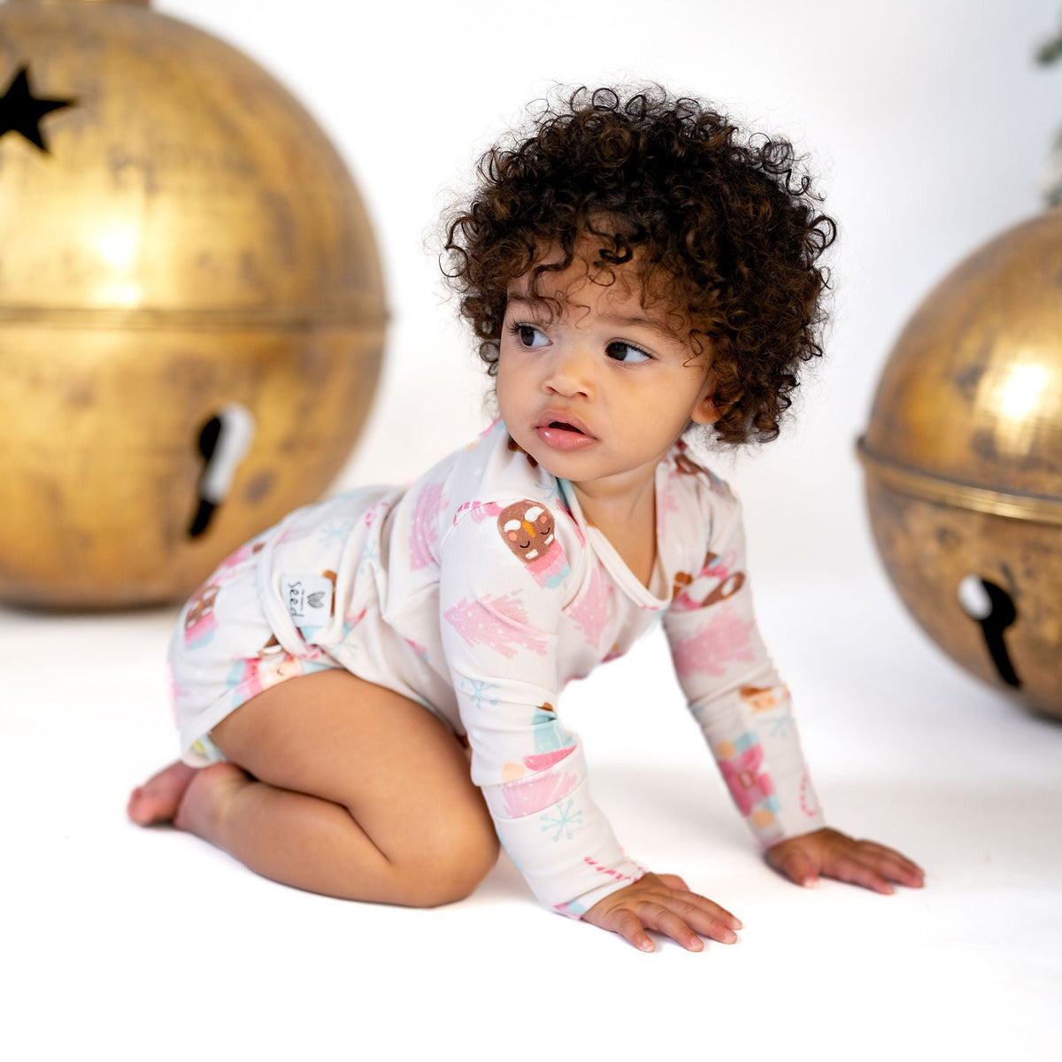 A baby with curly dark hair crawls on a white floor wearing a long-sleeve pajama top with a colorful holiday print. The background features two large, decorative gold jingle bells and a Christmas tree ornament.