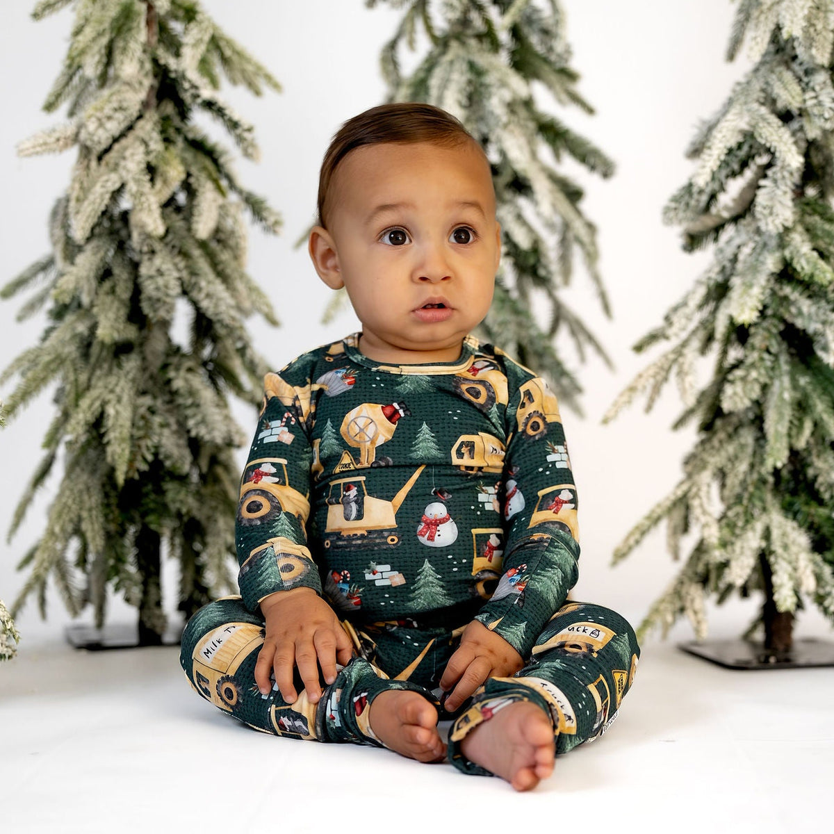 A baby sitting cross-legged on a white studio floor, wearing a dark green long-sleeve pajama set with a holiday construction print. The background features four artificial, snow-dusted Christmas trees.