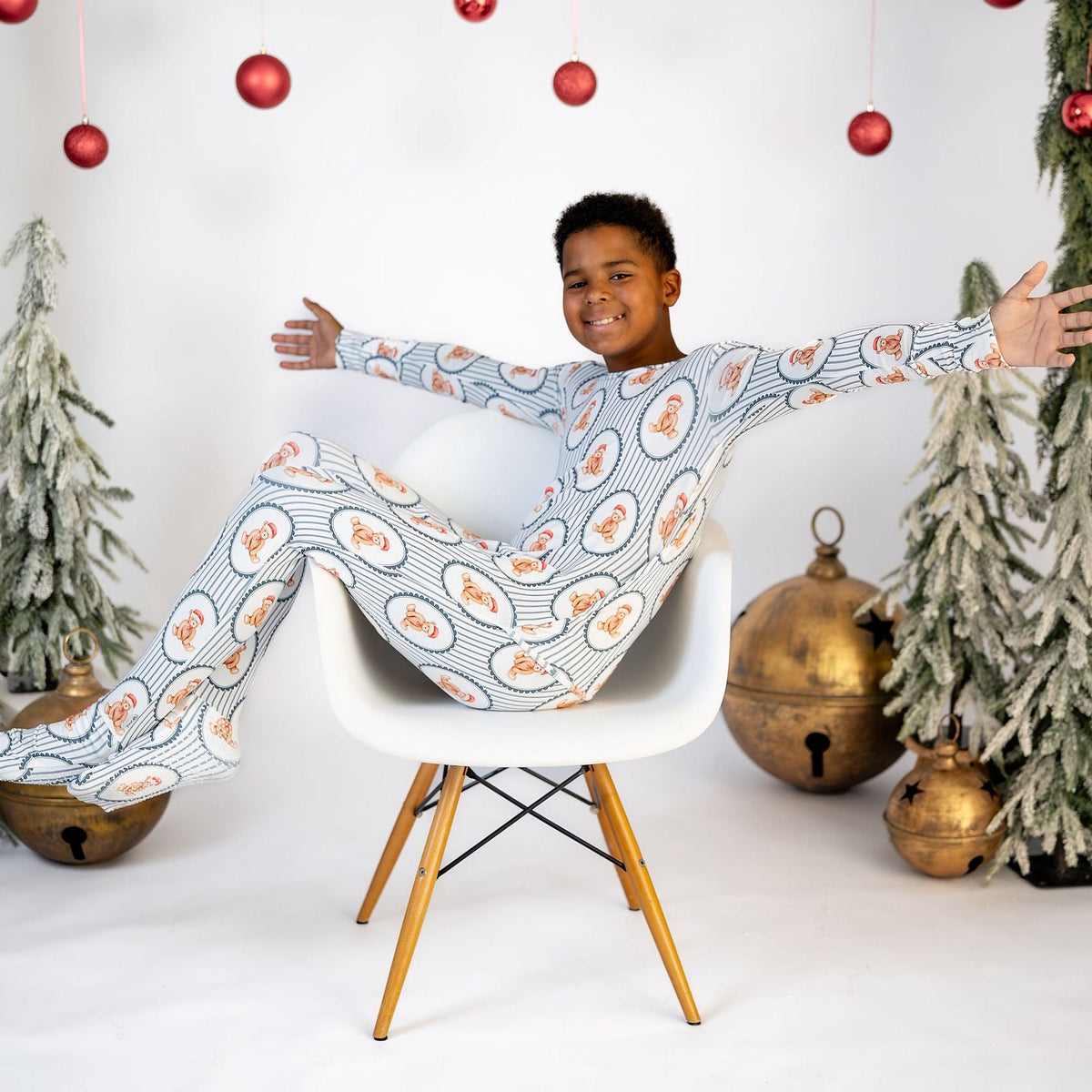A child wearing a festive Christmas-themed long-sleeve pajama set with a blue, white, and orange pattern, sitting on a white chair with arms outstretched in a decorated studio setting.