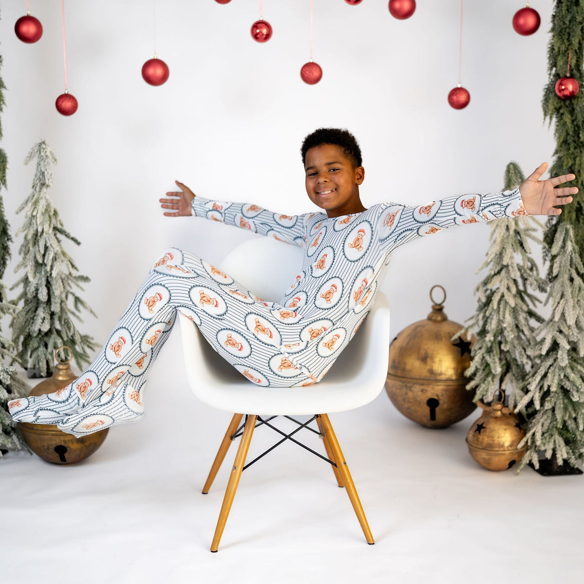 A child wearing a festive Christmas-themed long-sleeve pajama set with a blue, white, and orange pattern, sitting on a white chair with arms outstretched in a decorated studio setting.