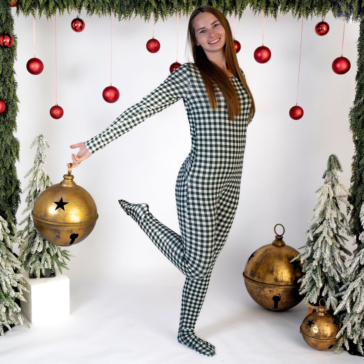A woman modeling a green and white gingham check long-sleeve footed jumpsuit, posing in a festive holiday studio setup with red ornaments, green garlands, and gold bells.
