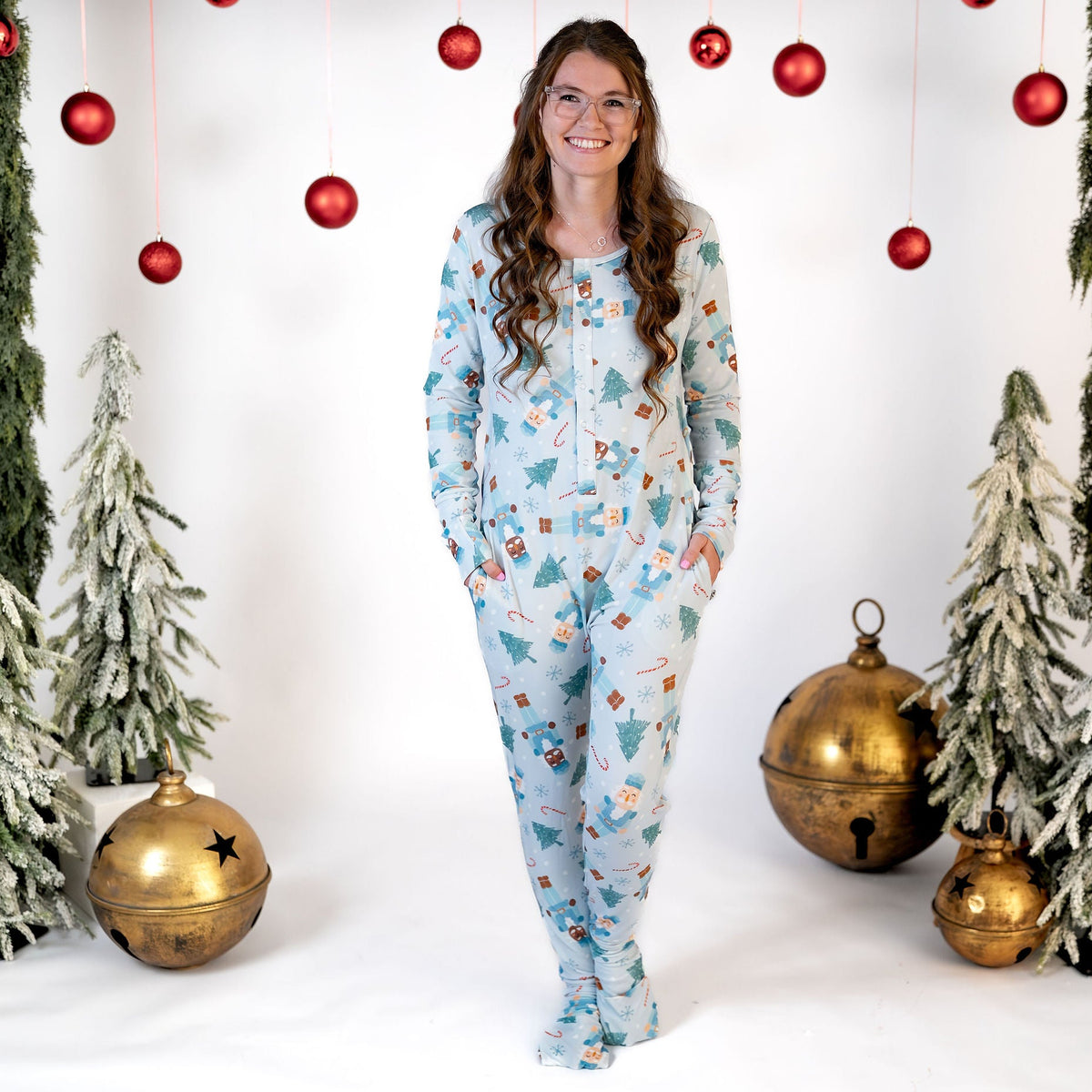 A woman stands in a studio wearing a light blue footed pajama set with a Christmas tree and gift print. The background is decorated with green garlands, red hanging ornaments, small frosted Christmas trees, and gold bells.