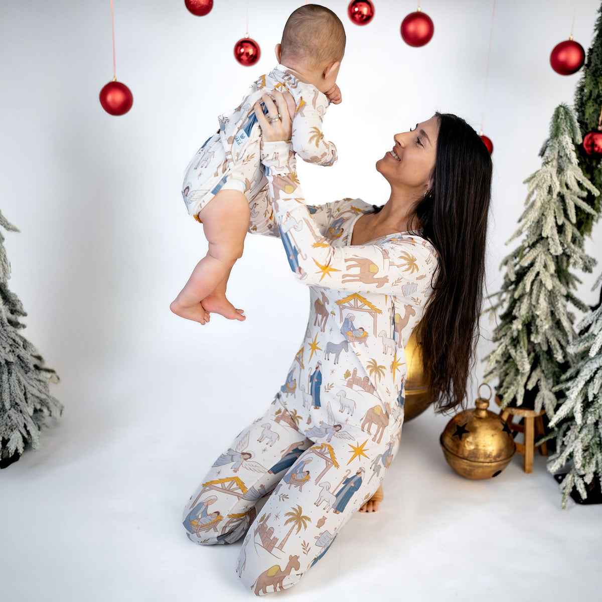 A woman and baby wearing matching animal-print pajamas pose together in a festive studio setting decorated with red hanging ornaments, artificial snowy trees, and a gold bell.