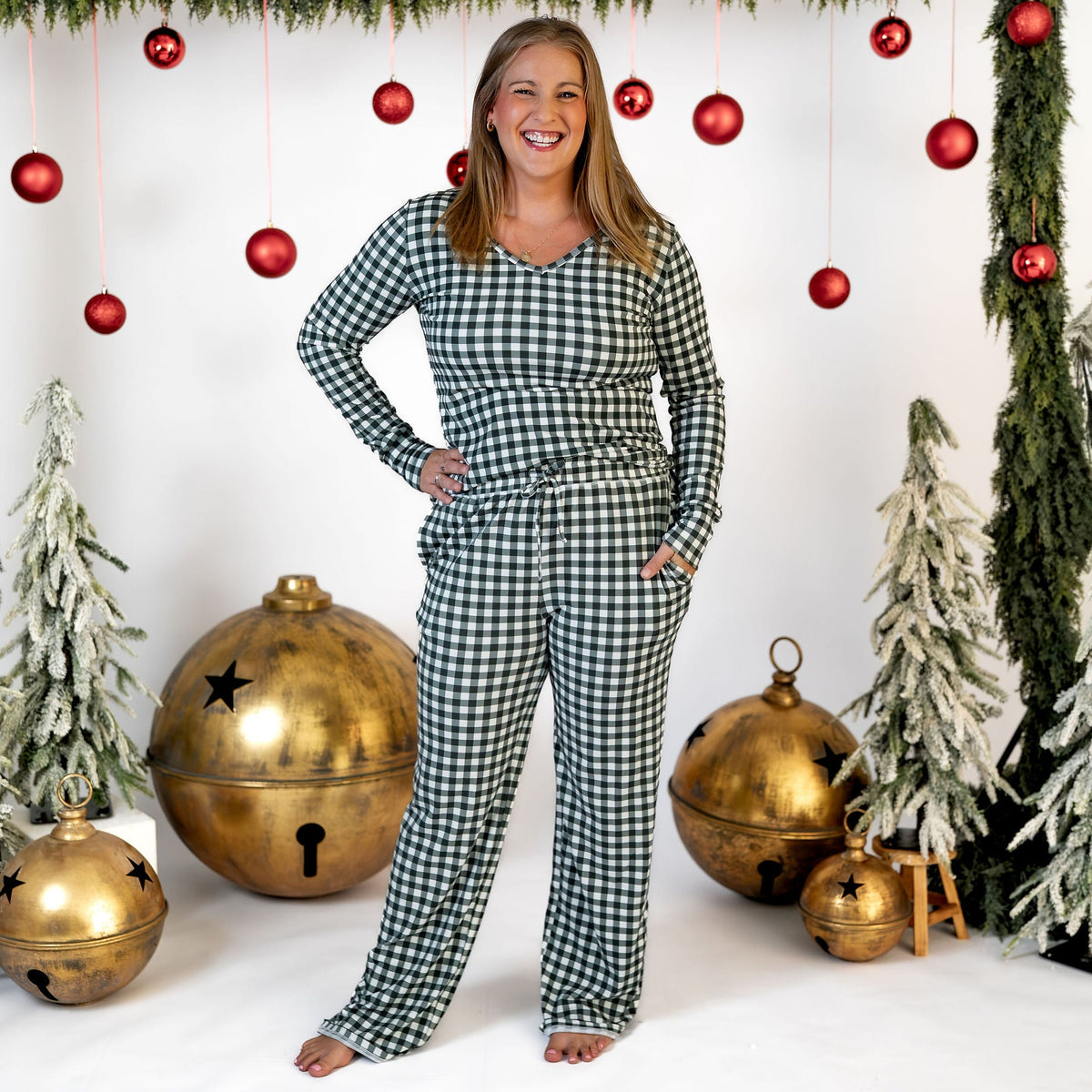 A woman stands barefoot in a holiday-themed studio setting wearing a matching green and white gingham check long-sleeve pajama set. The background features a white wall decorated with green garlands, red hanging ornaments, and large gold Christmas bells.