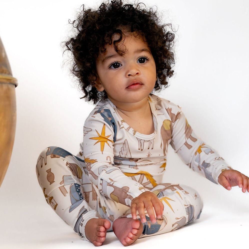 A baby with curly dark hair sits cross-legged on a white studio floor, wearing a long-sleeve one-piece pajama with a celestial print. The pajama has a cream base with gold, blue, and brown star and moon motifs.