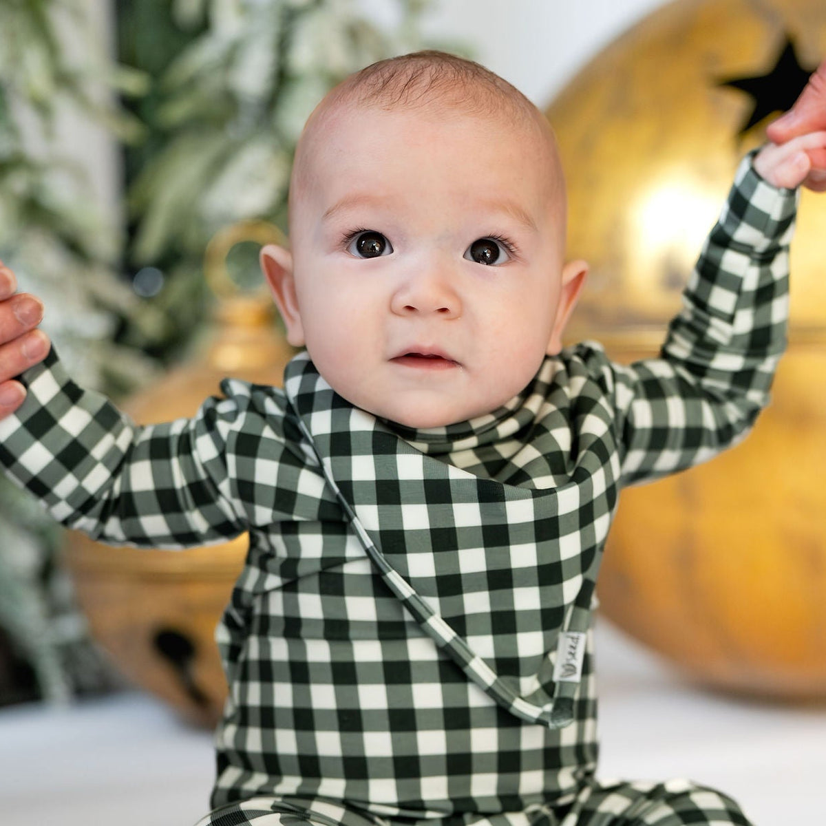 A baby wearing a green and white gingham footed sleeper sits on a white floor, holding hands with two adults' hands, with a gold bell and holiday tree in the background.