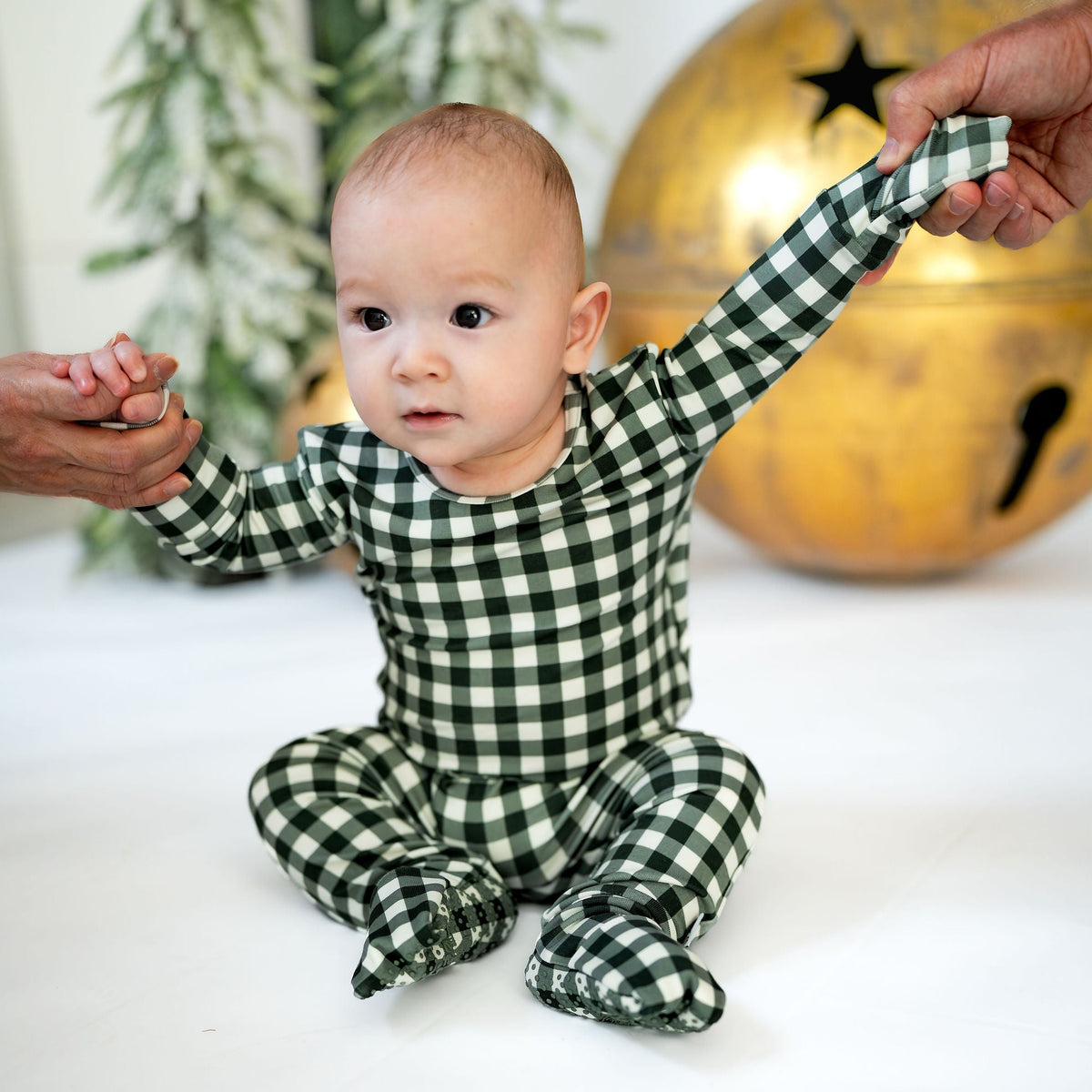 A baby wearing a green and white gingham footed sleeper sits on a white surface, holding hands with two adults, with a blurred festive background featuring a gold bell and greenery.