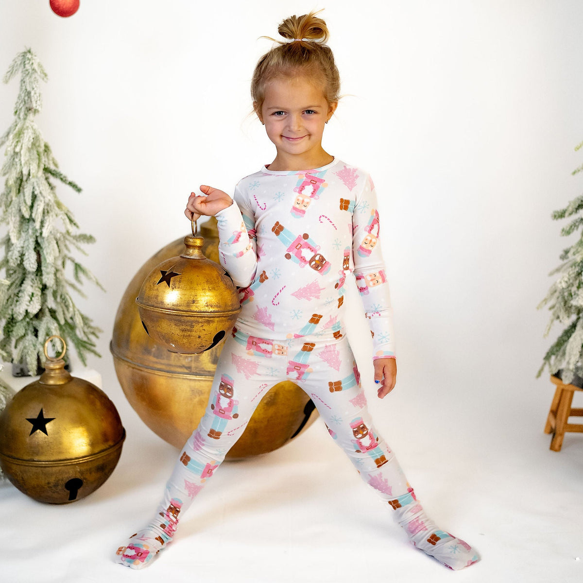 A young girl stands in a studio setting wearing a light pink and white long-sleeve pajama set with a Christmas ornament print. She is surrounded by holiday decorations, including a large gold bell, small frosted Christmas trees, and red hanging ornaments against a white background.
