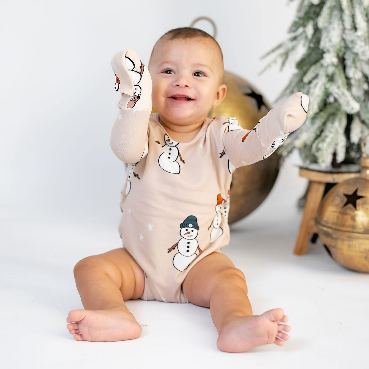 A baby sitting on a white studio floor wearing a light beige snowman print bodysuit, with a decorated Christmas tree and gold ornaments in the background.