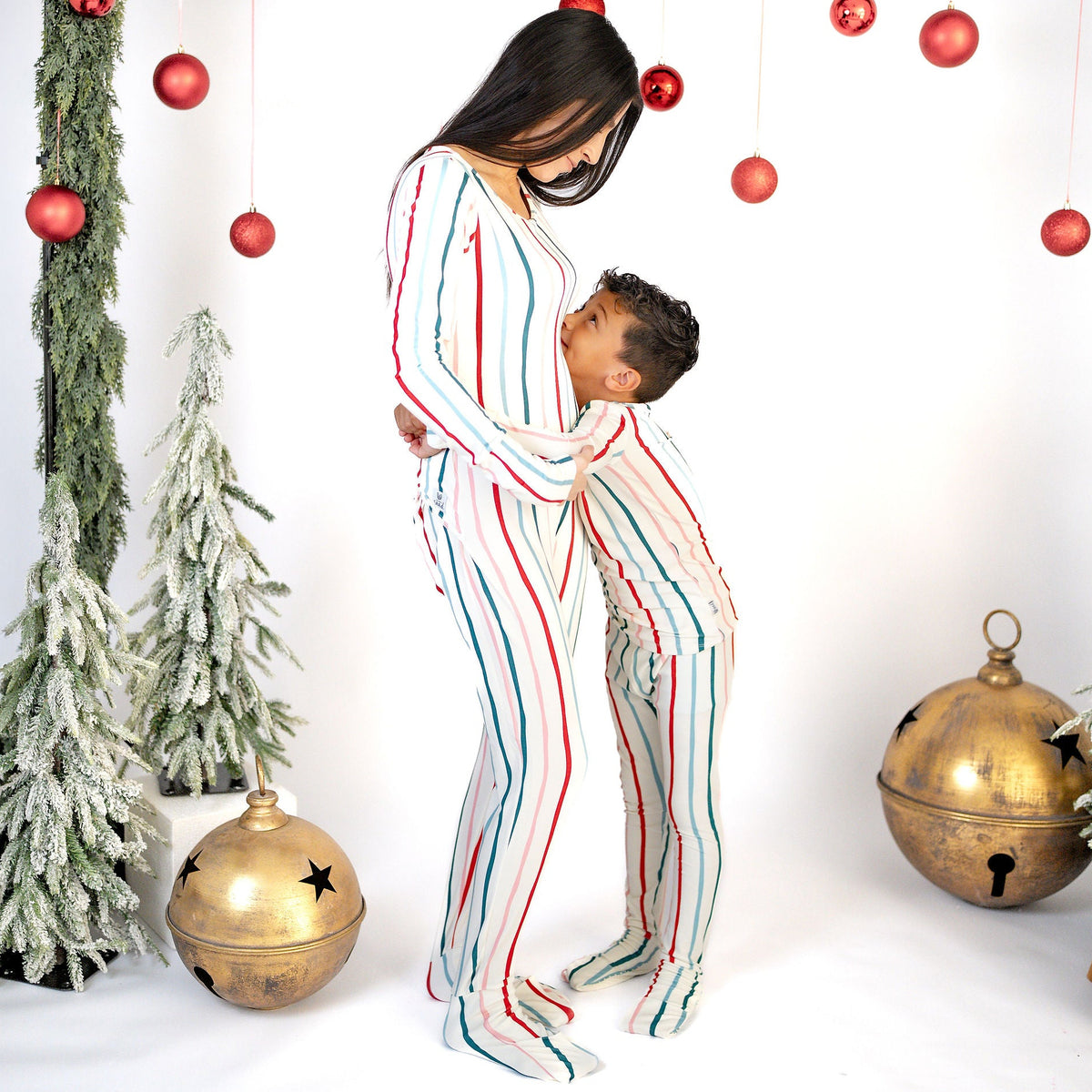 A mother and child wearing matching Christmas-themed pajamas with red, green, and blue vertical stripes on a white background, surrounded by holiday decorations including hanging red ornaments, a green garland, and gold bells.