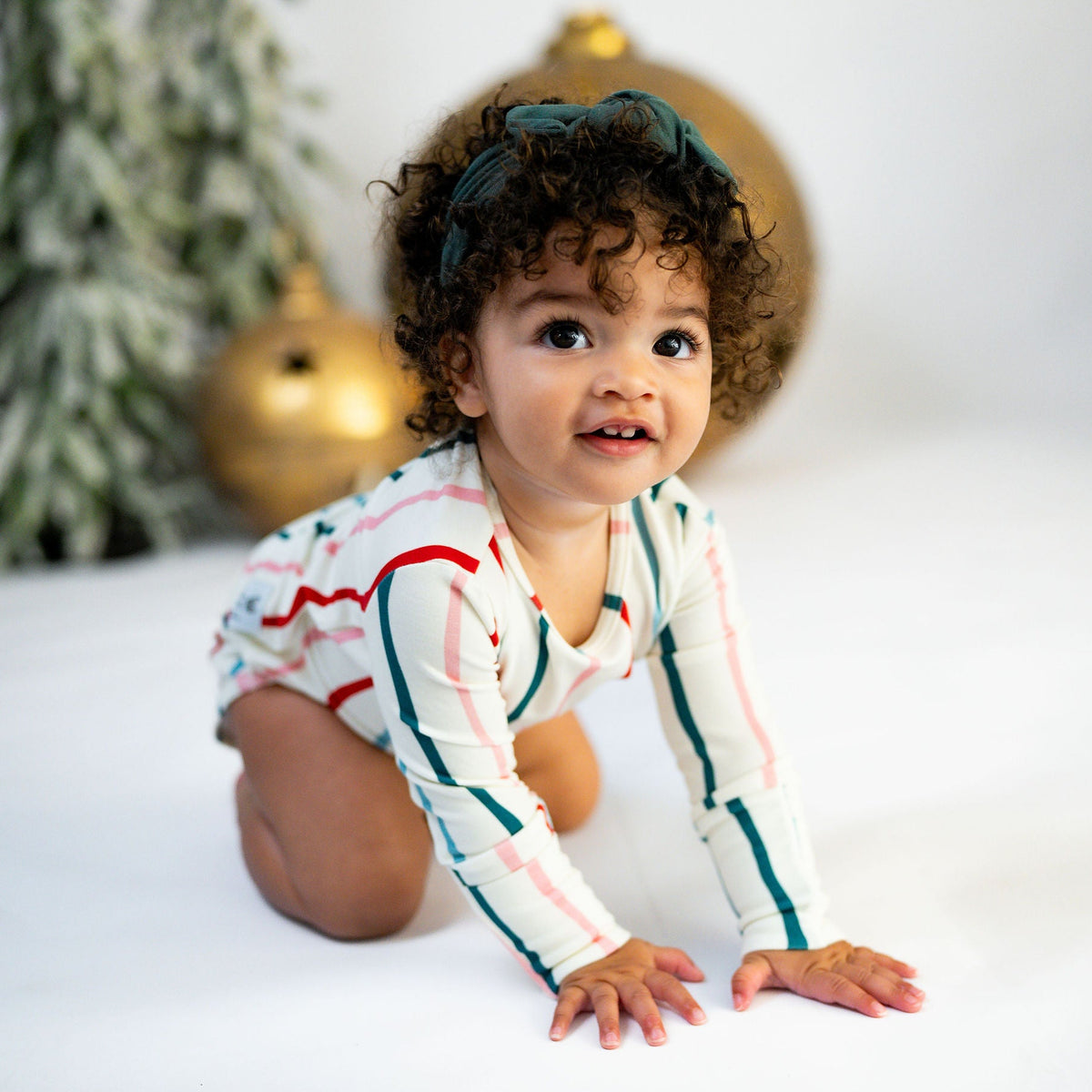 A baby wearing a long-sleeve striped romper and a dark green headband is crawling on a white studio floor with holiday decorations in the background.