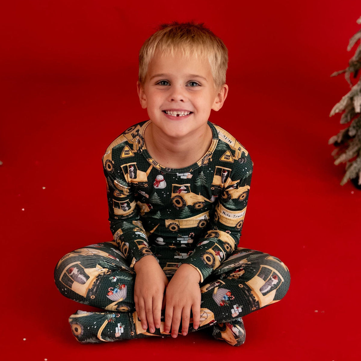 A young boy sits cross-legged on a bright red studio floor, wearing dark green and tan holiday-themed pajamas. Two decorative flocked Christmas trees frame the scene on either side against a solid red backdrop.