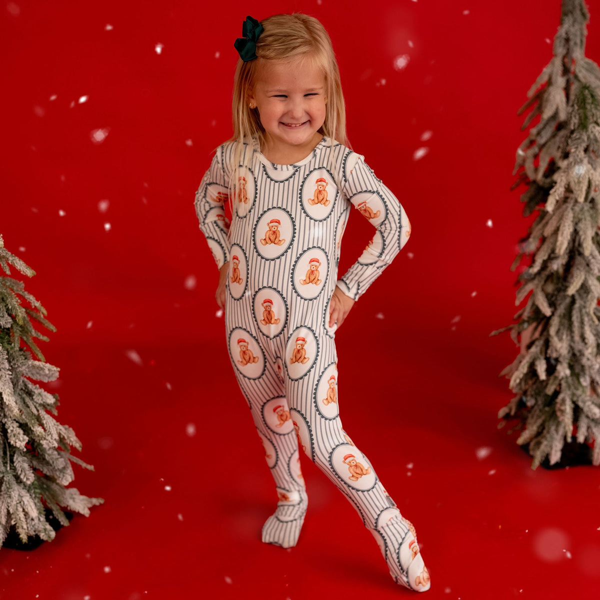 A young girl stands on a bright red studio background wearing a full-body knit one-piece pajama with a white base, gray vertical stripes, and orange gingerbread men motifs. Snowflake effects and small artificial trees decorate the scene.