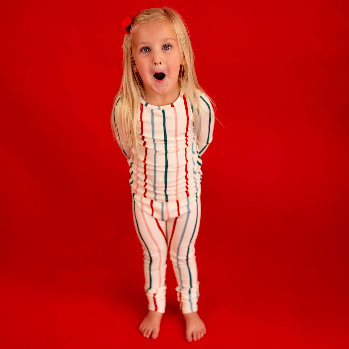 A young blonde girl with a red bow in her hair stands barefoot against a solid red background, wearing a white ruffled one-piece jumpsuit with vertical red and blue stripes.