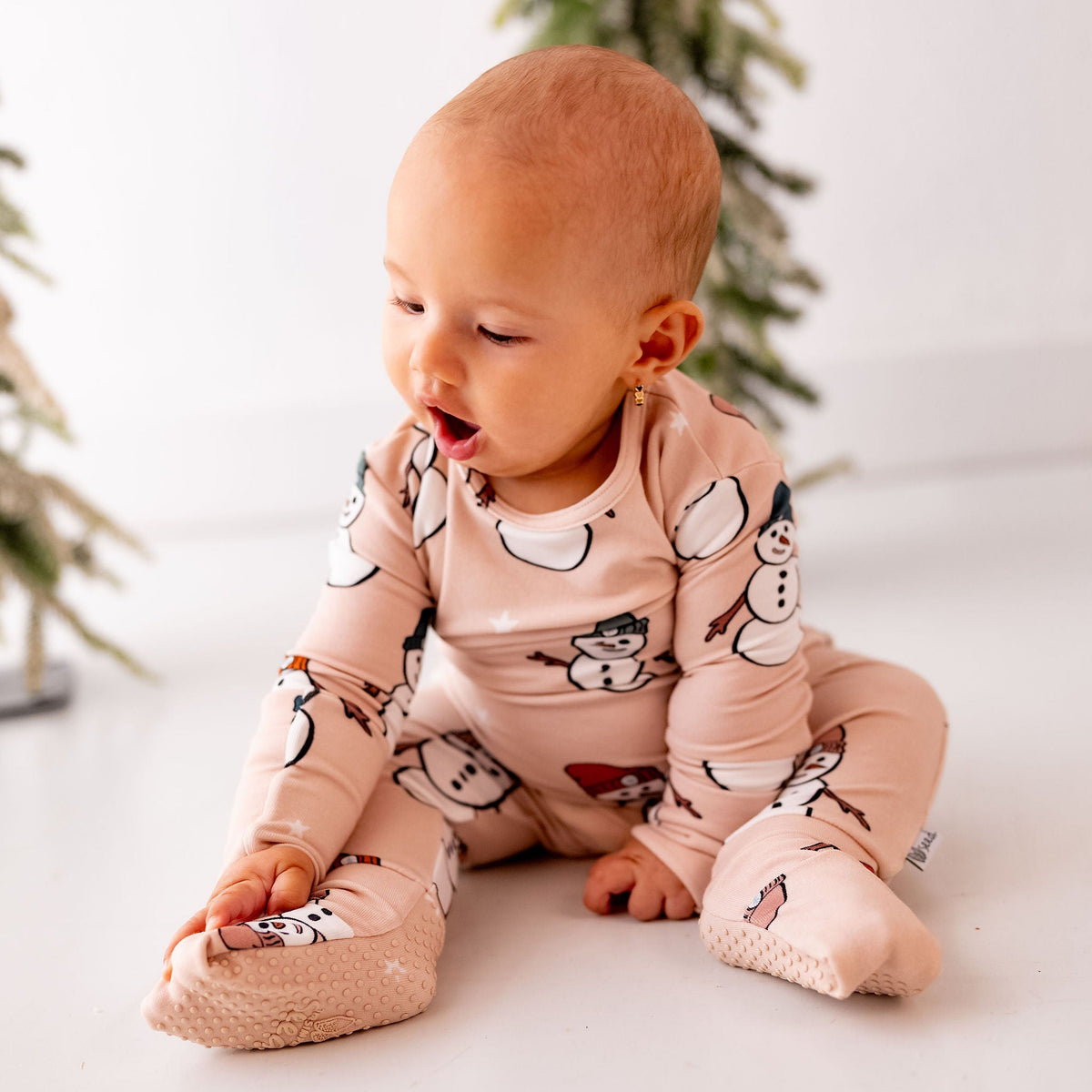 A baby sitting on a white floor wearing a light pink long-sleeve footed sleeper with a snowman and snowflake print, with blurred Christmas trees in the background.