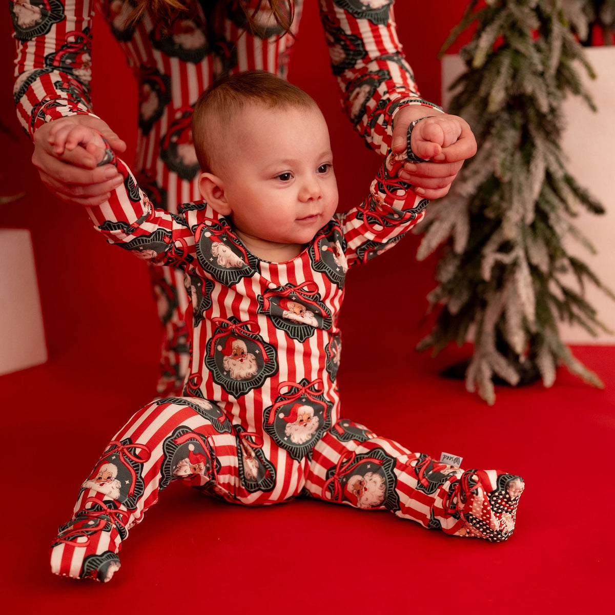 A baby wearing a red and white striped footed pajama with a black and white graphic print sits on a red floor while an adult holds the baby's hands. A decorated Christmas tree is visible in the background.