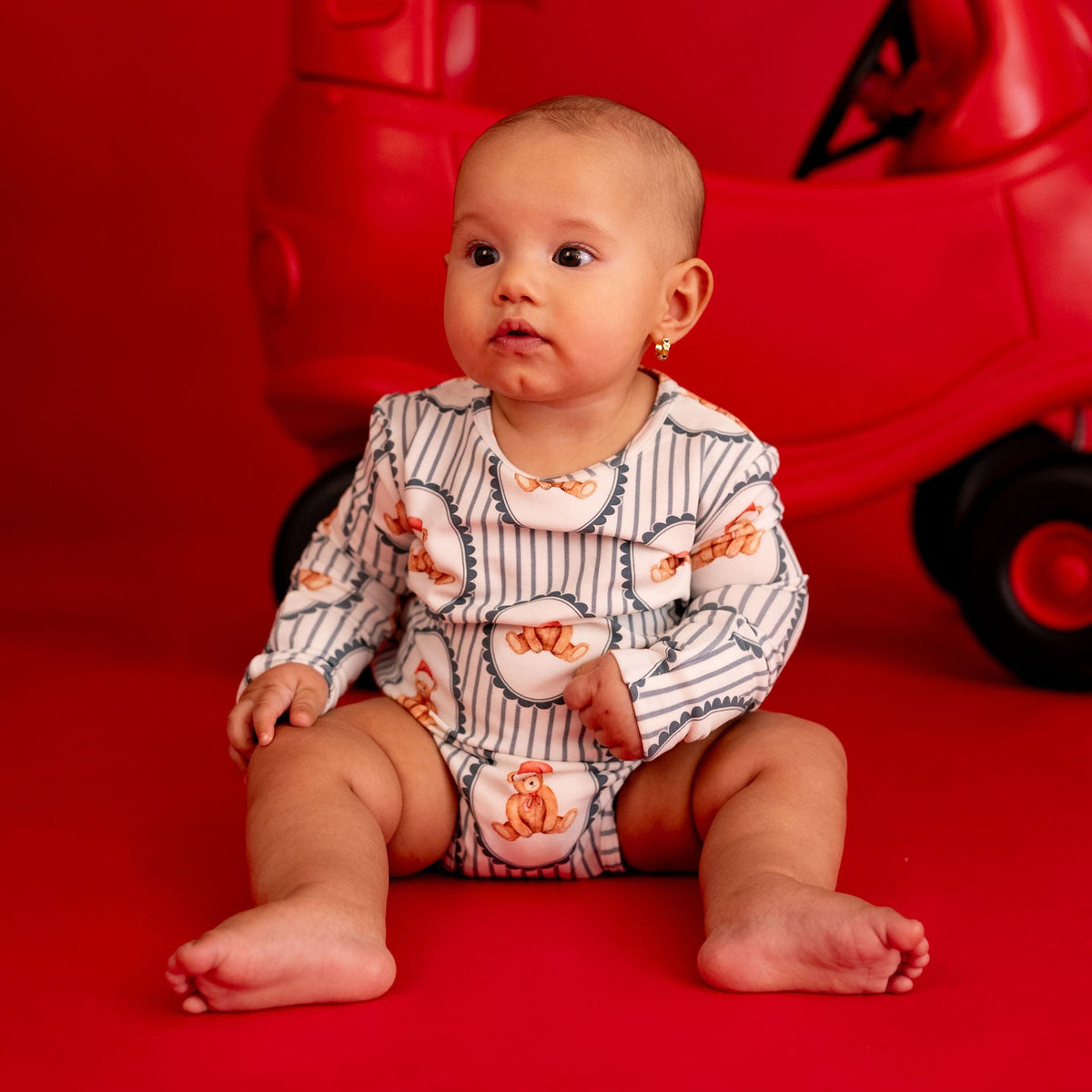 A baby sitting on a red studio floor in front of a red toy car backdrop, wearing a white long-sleeve romper with orange and navy pinstripes and a small orange animal print.