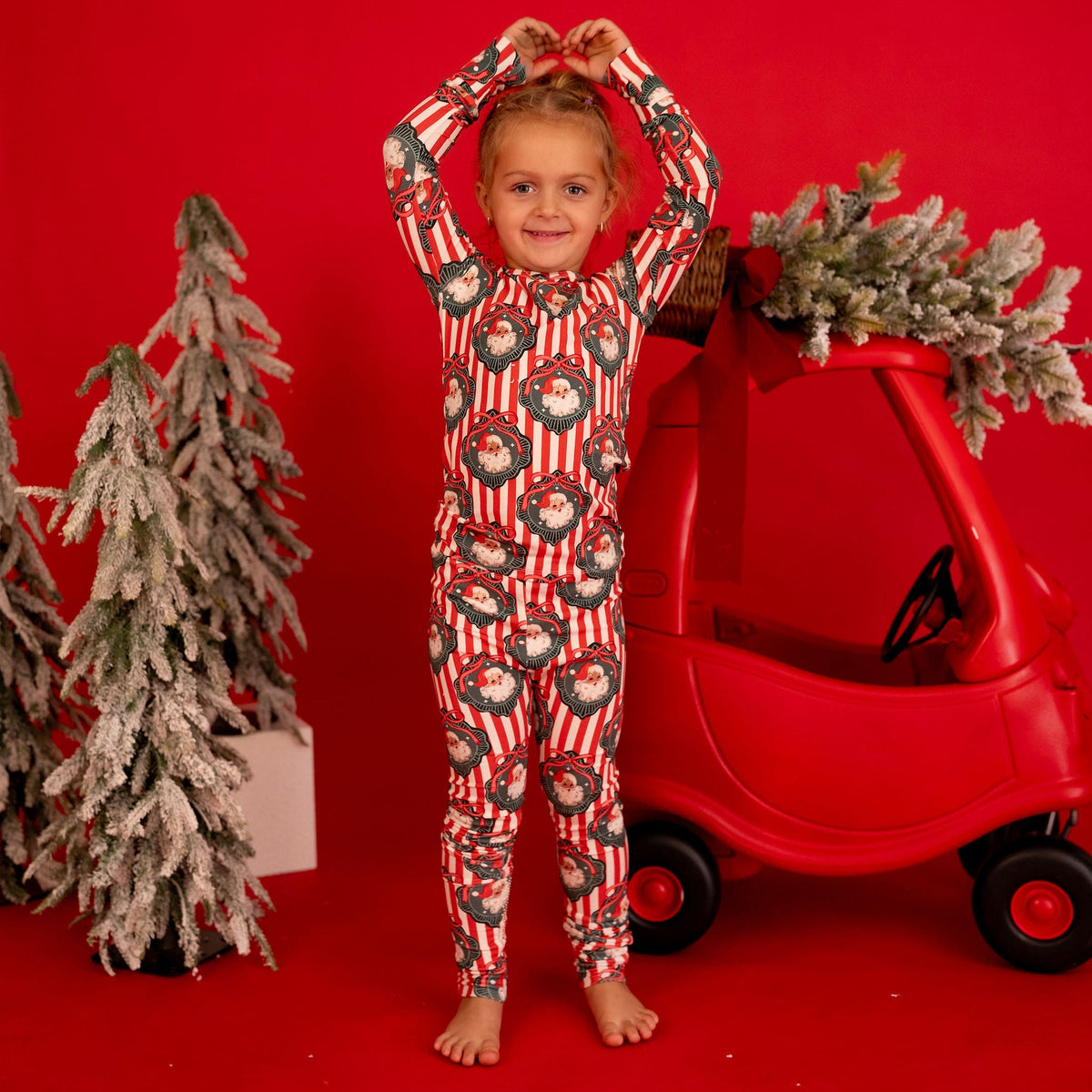 A young child stands barefoot on a bright red studio background, wearing a long-sleeve one-piece pajama with a holiday wreath pattern. The child is posed in front of a red backdrop with decorative flocked Christmas trees and a red toy car.