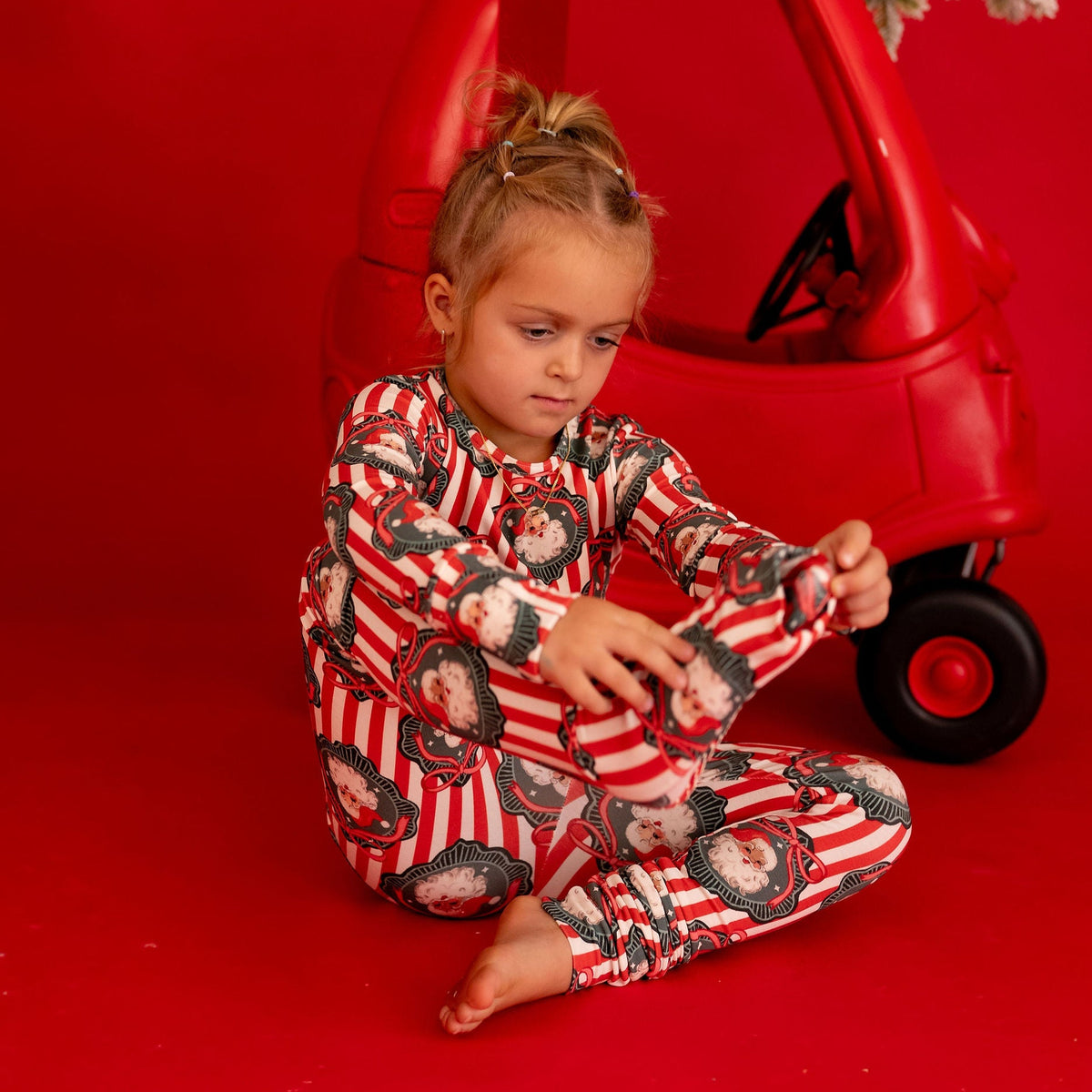 A young child sits on a red studio floor in front of a red toy car decorated with a small frosted evergreen branch. The child is wearing a red and white striped pajama set with a cartoon character print.