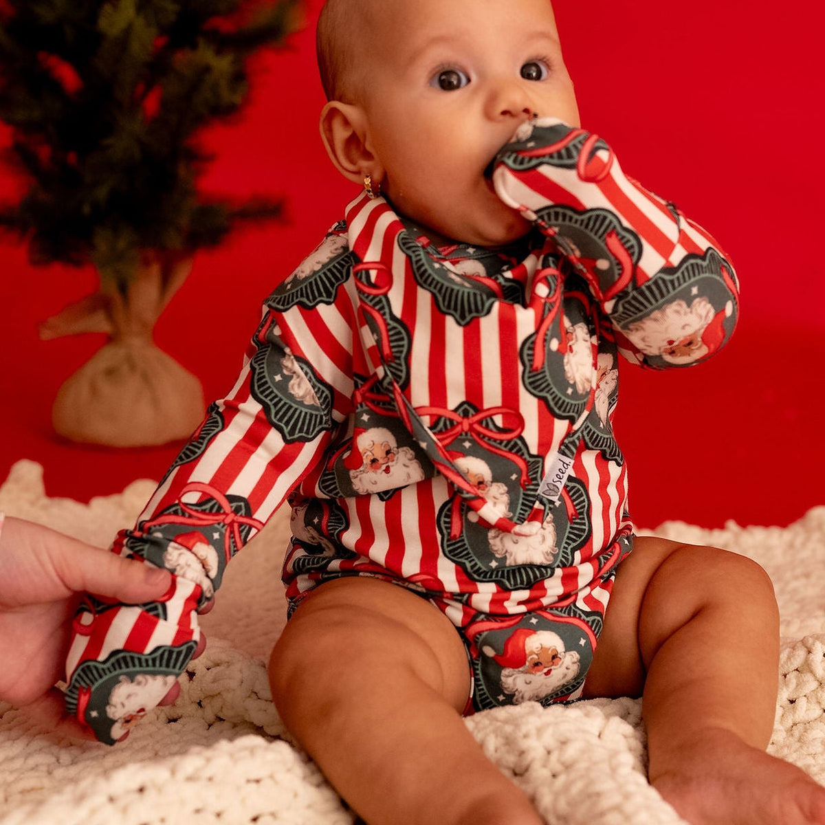 A baby wearing a red and white striped long-sleeve bodysuit with a dark green holiday tree print sits on a cream knit blanket against a solid red background, with a small artificial Christmas tree and a hand adjusting the blanket.
