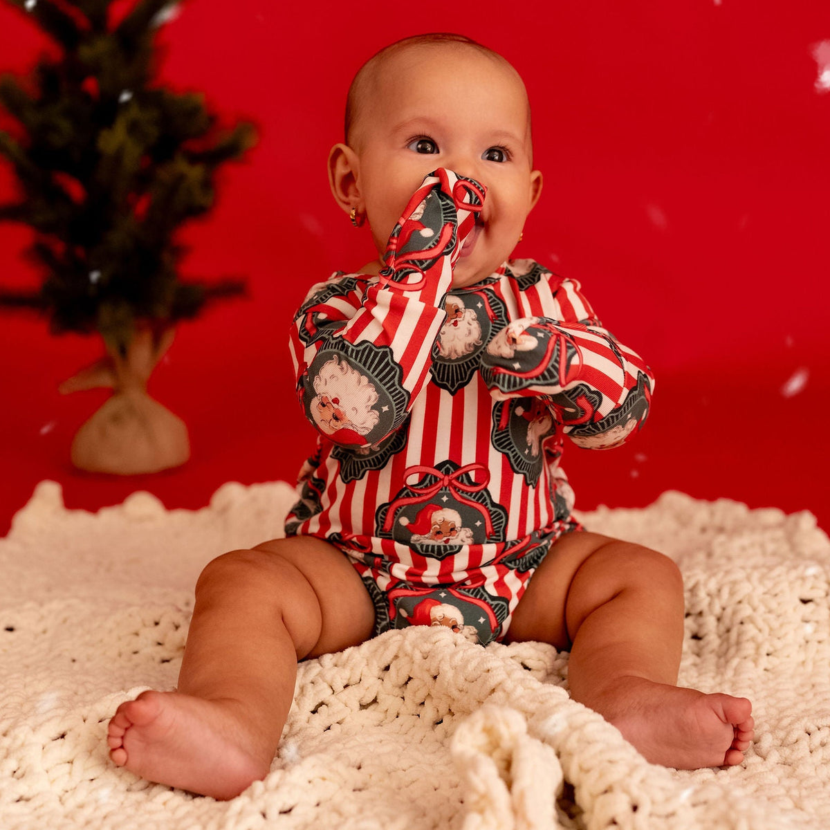 A baby sitting on a cream knit blanket against a bright red backdrop, wearing a red and white striped one-piece outfit with a cartoon bear print. A small artificial tree stands to the left, and white bokeh snow-like dots float in the scene.