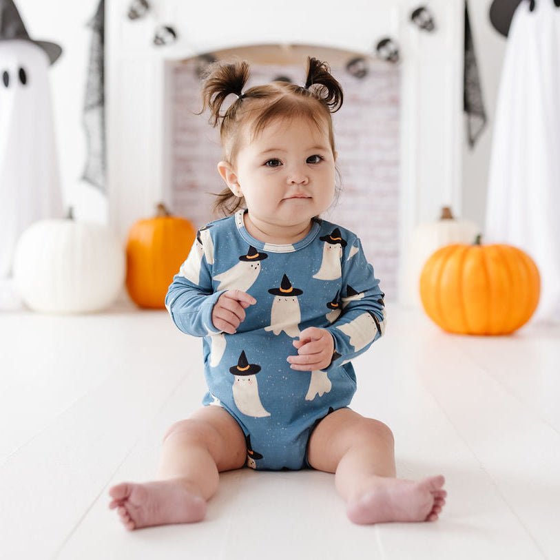 A baby sitting on a white floor wearing a blue long-sleeve romper with a penguin pattern, surrounded by Halloween decorations including pumpkins, ghost cutouts, and a fireplace backdrop.