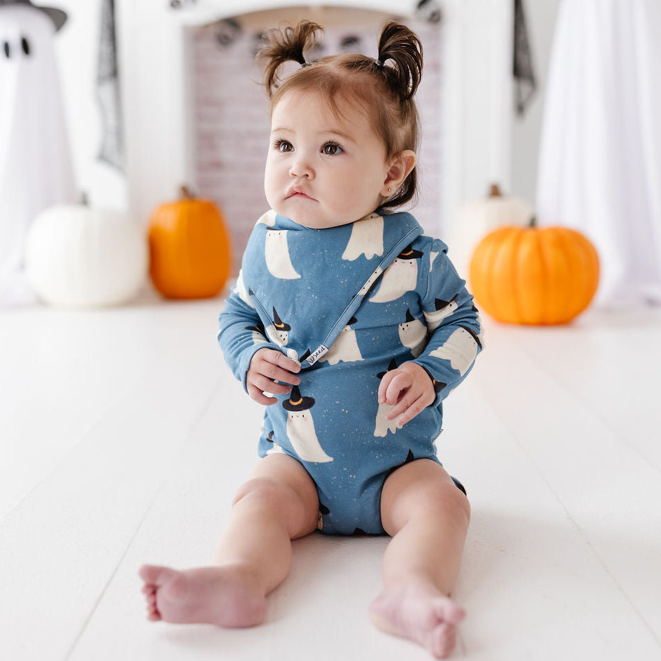 A baby wearing a blue long-sleeve romper with a white penguin print sits on a white floor in a Halloween-themed room with pumpkins and ghost decorations.