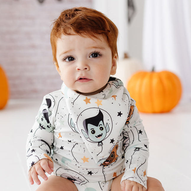 A baby with red hair sits on a white floor wearing a long-sleeve white space-themed bodysuit with cartoon characters and stars. The background features soft-focus orange pumpkins and a white curtain.