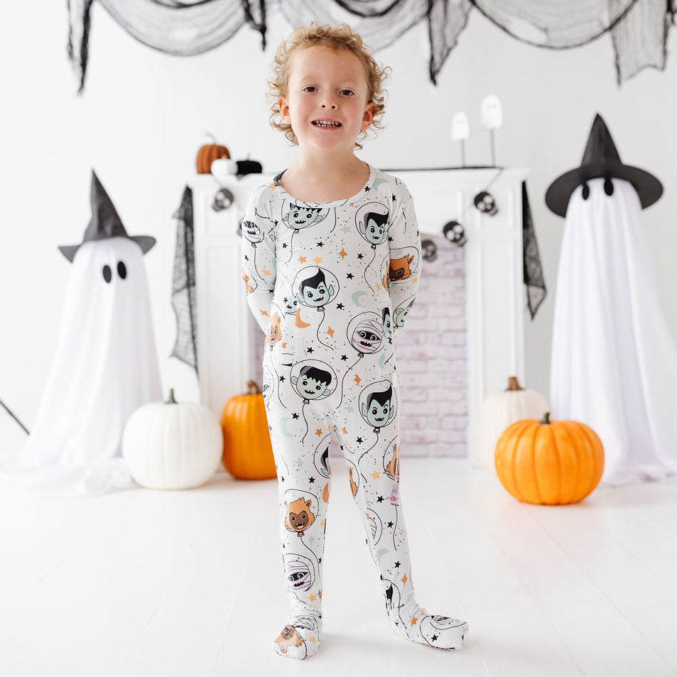 A young child stands in a Halloween-themed room wearing a white footed pajama with a black cat astronaut print. The background features a white fireplace mantel decorated with black spiderwebs, ghost figures in white sheets, orange pumpkins, and black spider decorations.