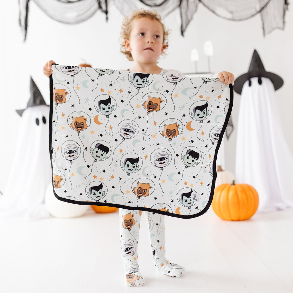 A young child stands holding up a white blanket or sheet with a repeating Halloween vampire balloon pattern, in a decorated Halloween-themed room with pumpkins and ghost props.