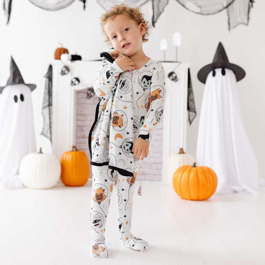 A toddler stands in a Halloween-themed room wearing a long-sleeve footed pajama set with a white base and a cartoon spider pattern. The room features a white fireplace mantel decorated with black spiderwebs, orange pumpkins, and ghost props.