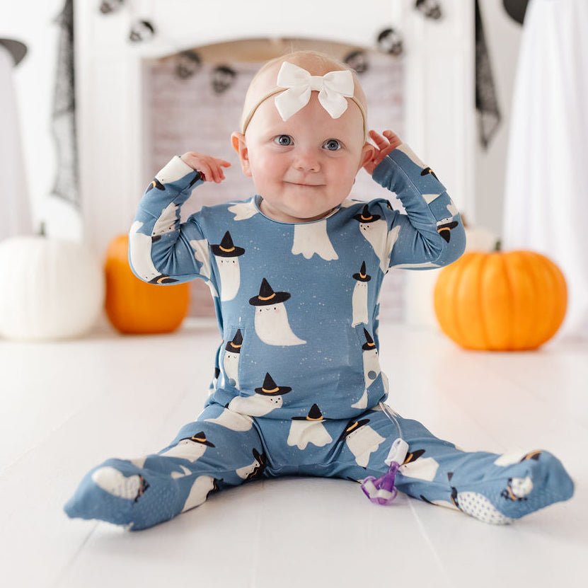 A baby sitting on a white floor wearing a blue footed onesie with a ghost pattern, styled in a Halloween-themed room with pumpkins and ghost decorations.