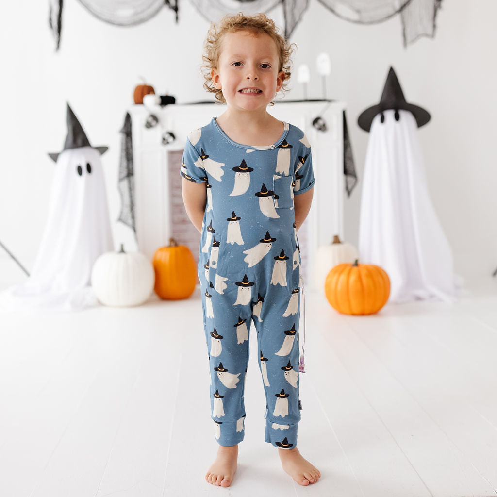 A young child stands barefoot in a white room decorated for Halloween, wearing a blue short-sleeve one-piece pajama with a repeating ghost pattern. Ghosts are white with black pointed hats, and pumpkins are placed on the floor around the child.