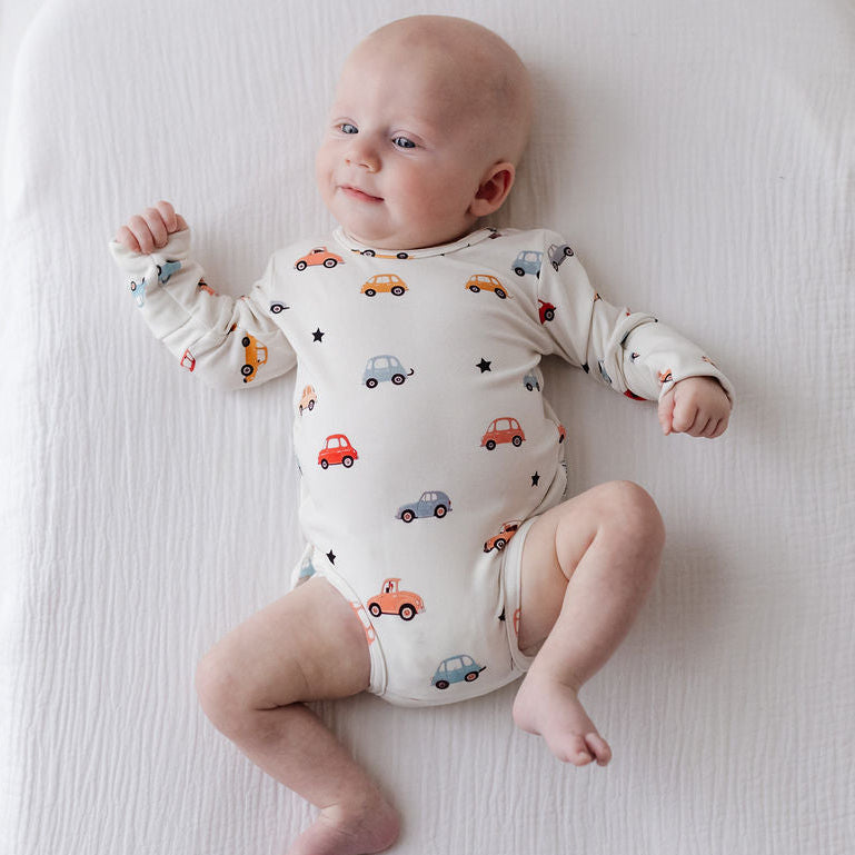 A baby lying on a white muslin changing pad, wearing a cream-colored long-sleeve bodysuit with a colorful pattern of small cars.