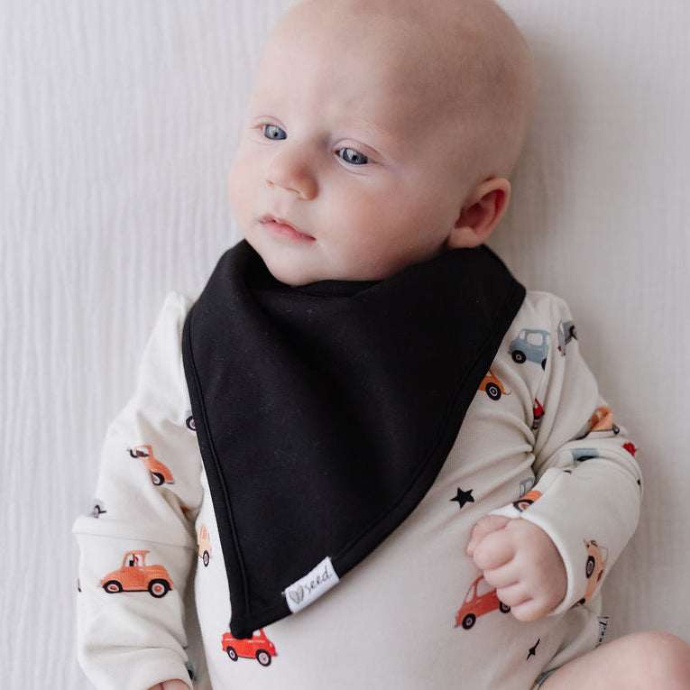 A baby lying on a white textured blanket, wearing a cream-colored long-sleeve bodysuit with a car pattern and a black triangular bib.