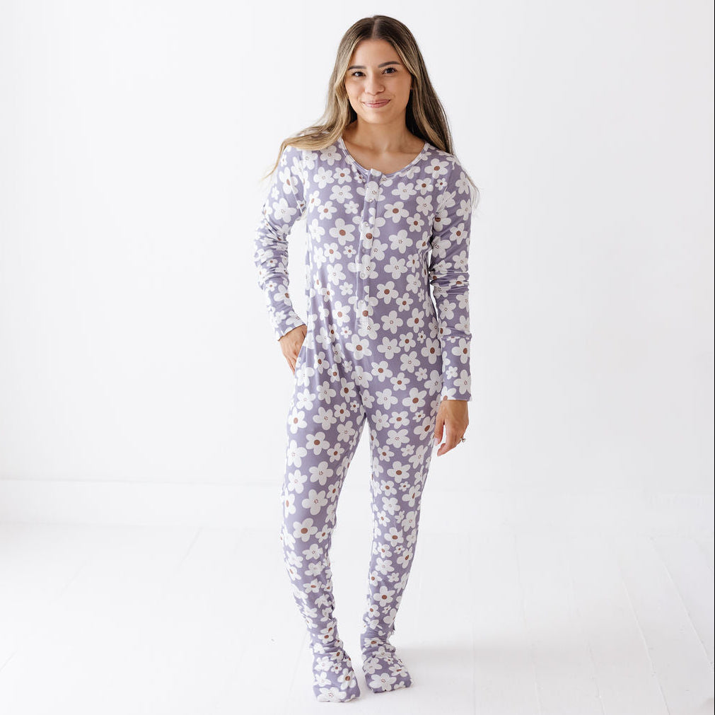 A woman modeling a one-piece footed pajama top with long sleeves and an allover daisy floral print in lavender, white, and yellow, set against a plain white studio background.