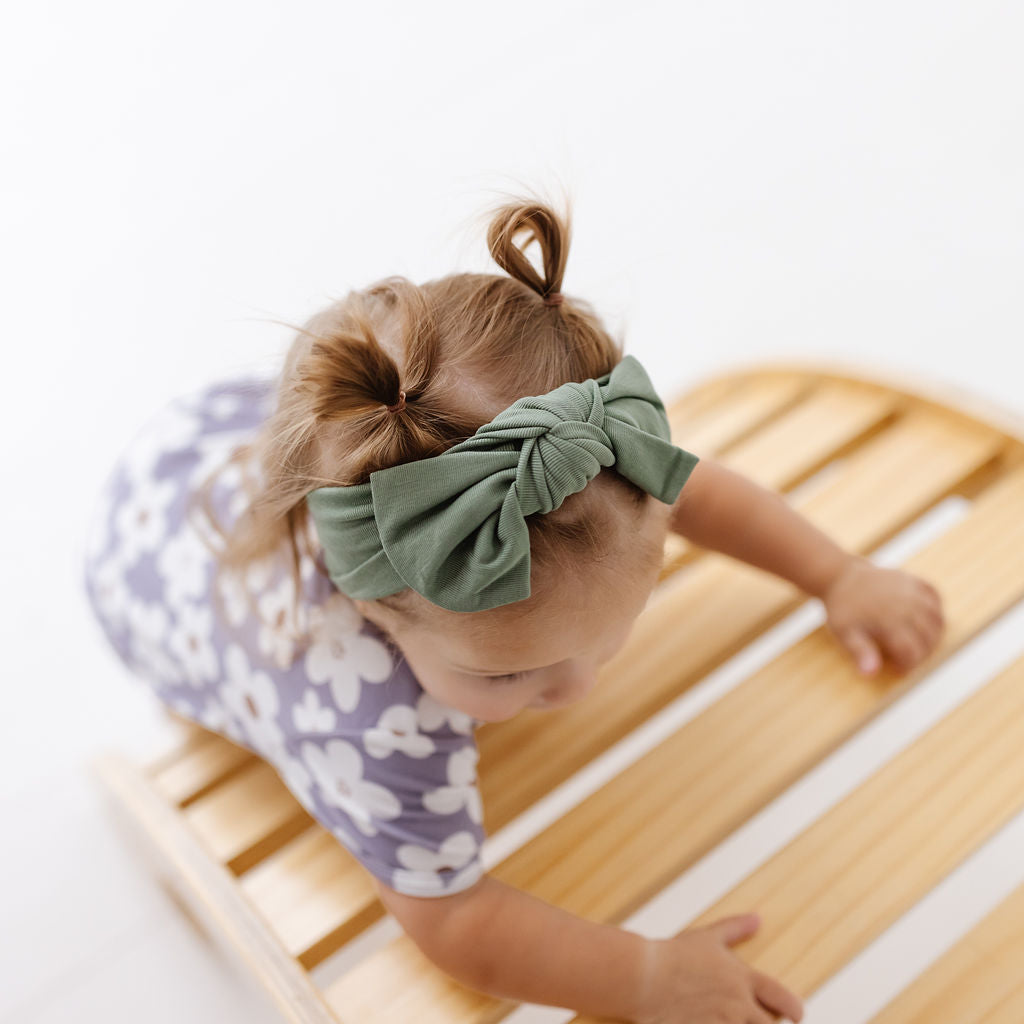 A baby girl crawling on a wooden slatted bench, wearing a sage green knotted fabric headband and a purple floral short-sleeve romper.