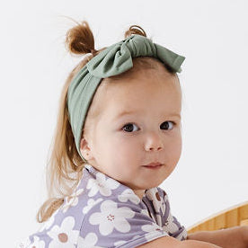 A toddler girl in a floral print romper and green headband is climbing on a light natural wood arch play structure in a bright white studio setting.