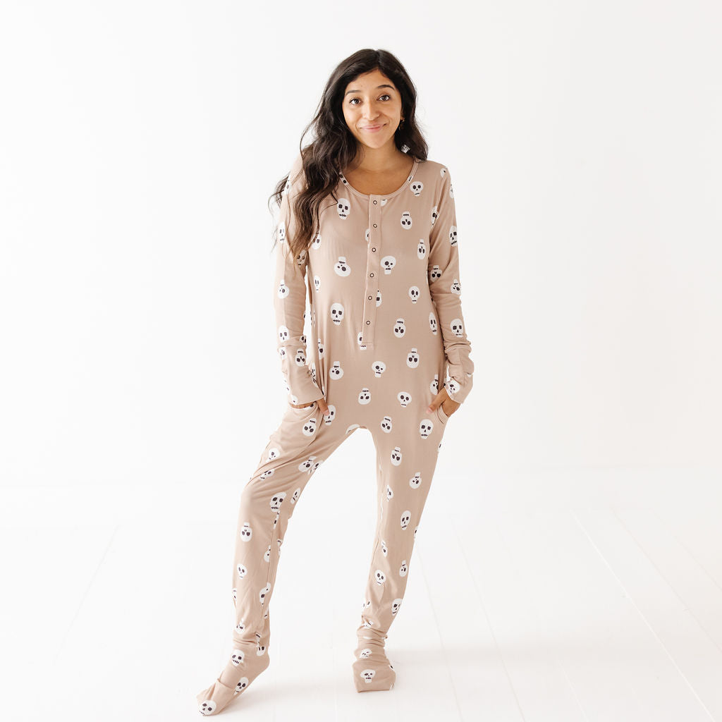 A woman modeling a beige footed pajama one-piece with an all-over white skull pattern, standing against a clean white studio background.