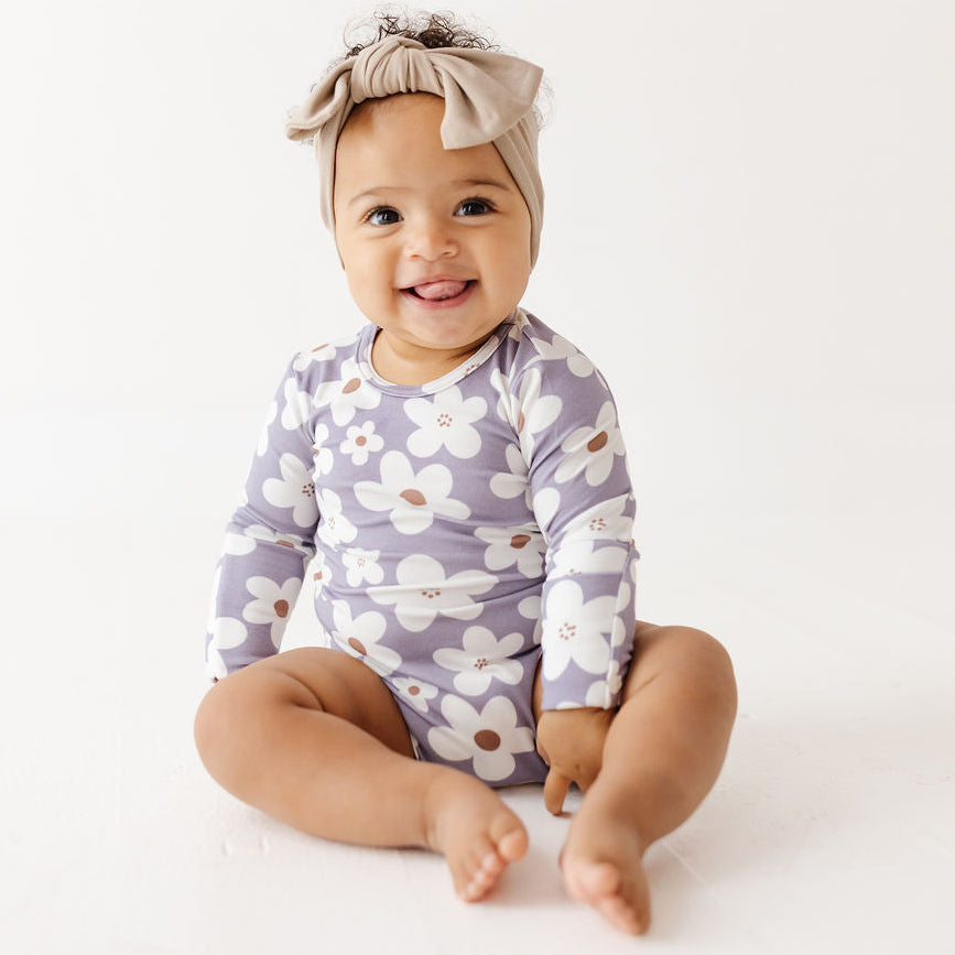 A smiling baby sitting on a white studio floor, wearing a long-sleeve floral romper in lavender and white with a beige bow headband.