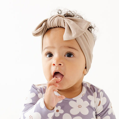 A baby sitting on a white floor wearing a long-sleeve floral romper in lavender and white, along with a beige bow headband.